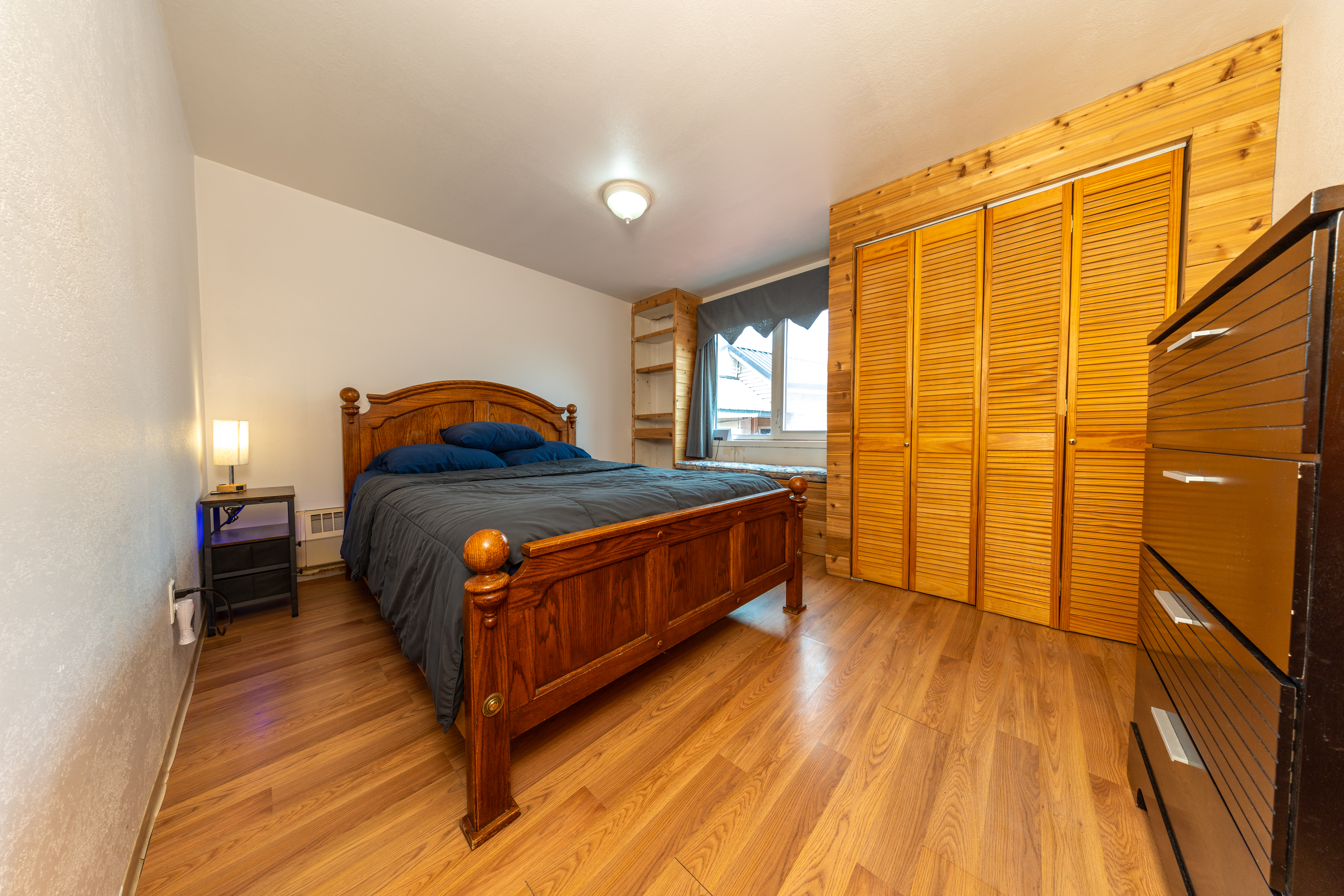 Bedroom with wooden bed, dresser, and closet doors, featuring wood floors and window with natural light
